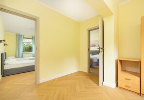 A room with a wooden floor, a bed with white bedding, a wooden cabinet with drawers, and a door leading to another room. - Ale Fale Trzęsacz Apartment #1 (Trzęsacz)