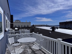 A rooftop terrace with a metal table and chairs, a white railing, and a view of buildings in the distance. - State + Main Condo | Downtown Luxury Studio (Harbor Springs)