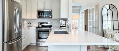 A modern kitchen with stainless steel appliances, a central island with stools, and a view to the living area.