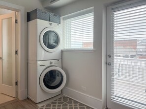 A laundry room with two stacked washing machines, a window with blinds, and a door. - State + Main Condo | Downtown Luxury Studio (Harbor Springs)