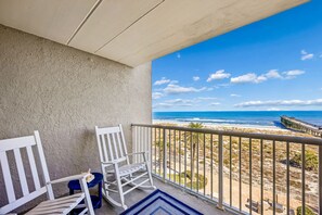 A balcony with two white rocking chairs and a blue rug, overlooking a beach with a pier. - 303 Atlantis 2 Bedroom Condo (Fernandina Beach)