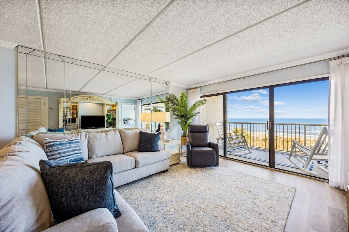 A living room with a sofa, armchairs, a TV, and a view of the beach through a sliding glass door.