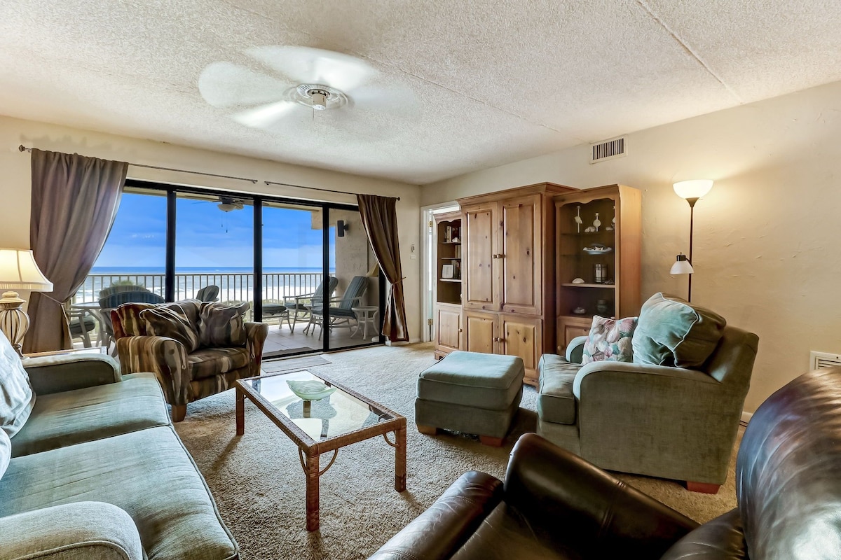A spacious living room with a large window, a ceiling fan, and a view of the sea.