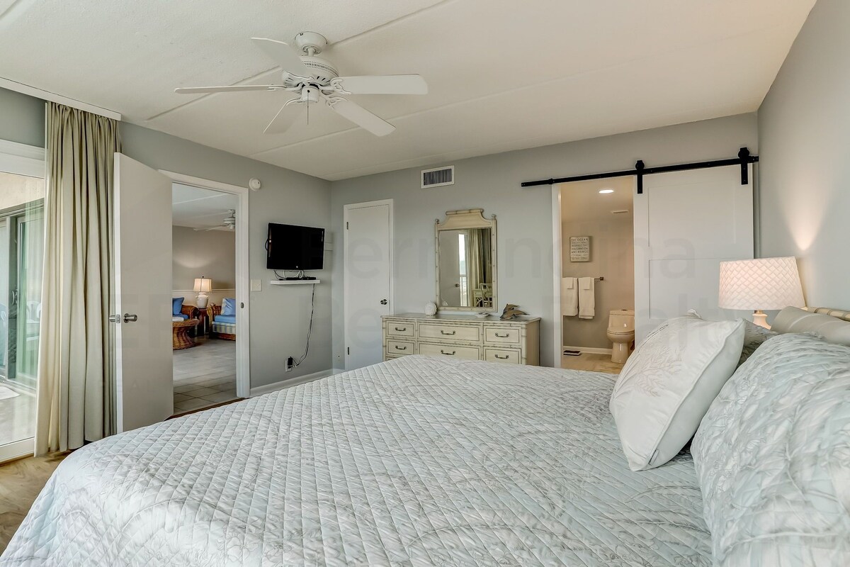 A bedroom with a large bed, a dresser, a ceiling fan, a television, and a sliding door leading to another room.