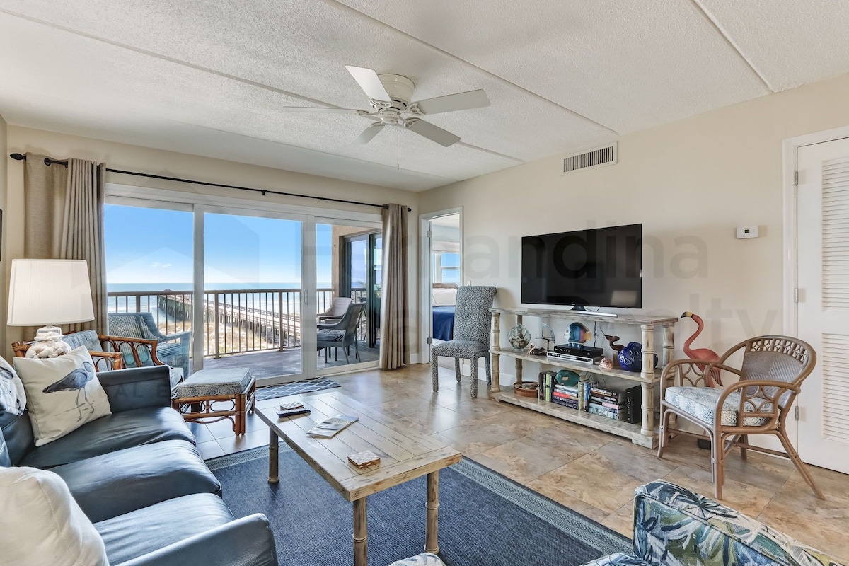 A spacious living room with a large TV, comfortable seating, a wooden coffee table, and a view of the ocean through large windows.