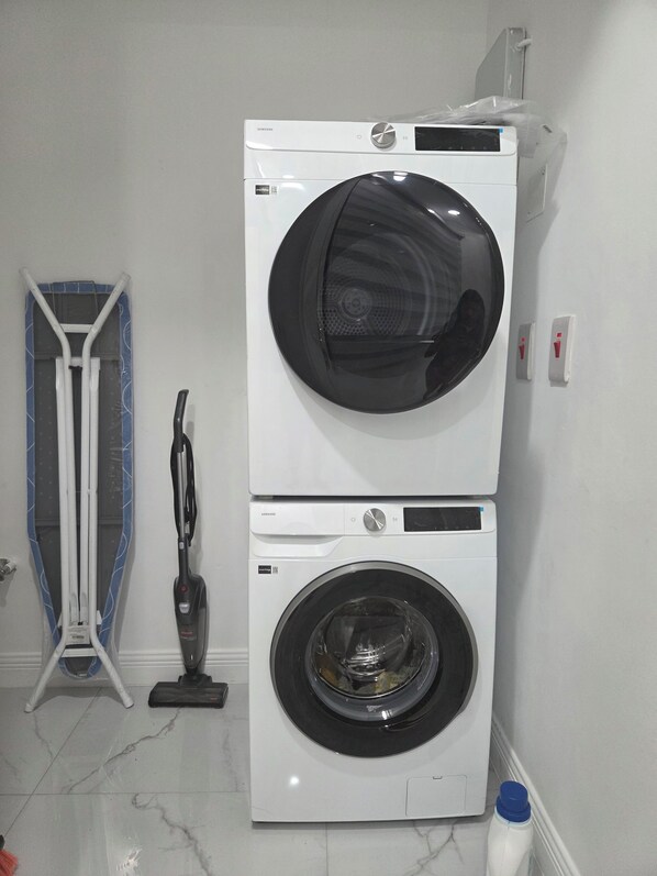 A stacked washer and dryer unit in a room with a tiled floor and a vacuum cleaner. - Charisma Airbnb (Kingston)
