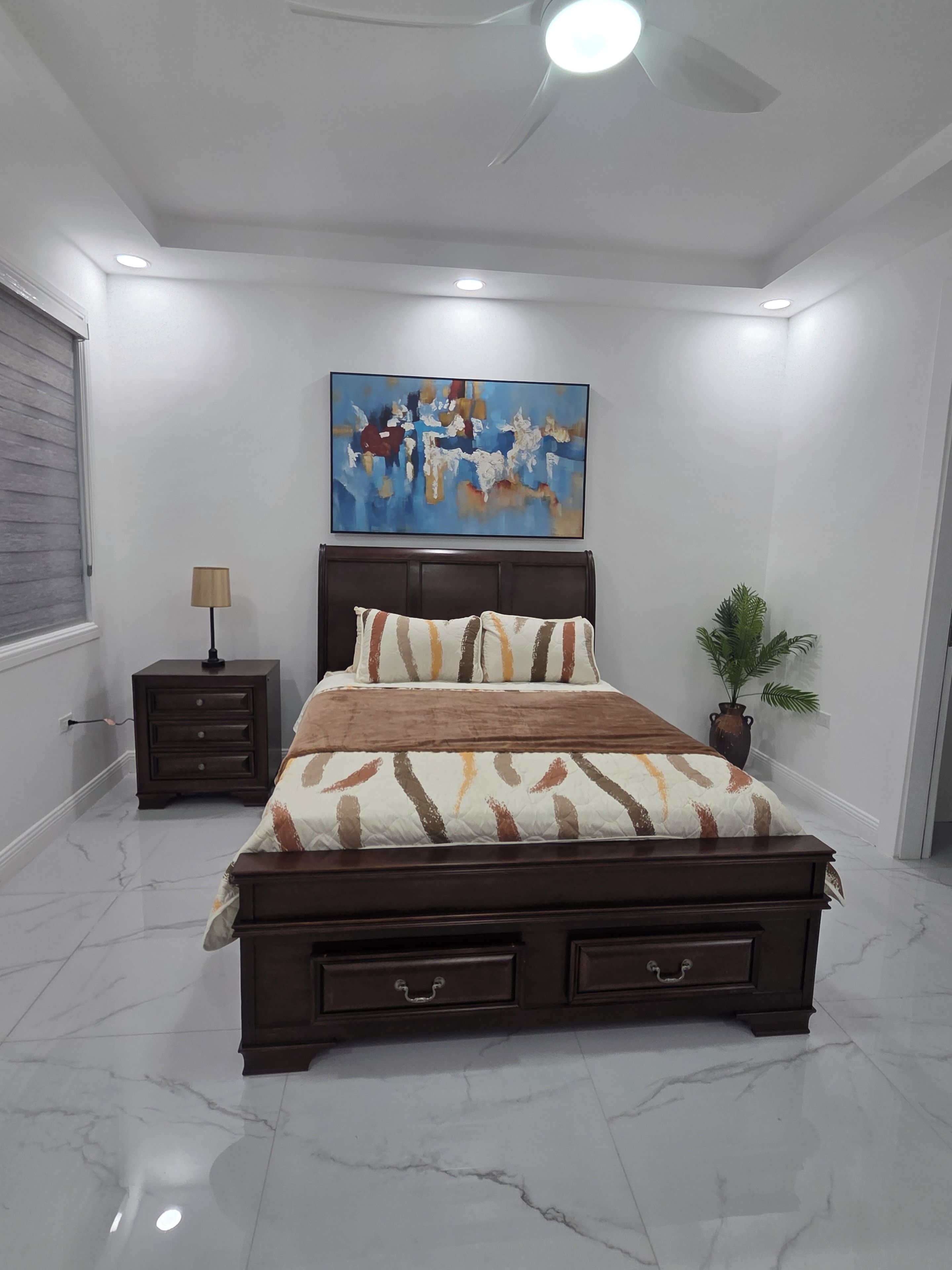 A bedroom with a bed, a nightstand, a ceiling fan, a painting, and a potted plant.