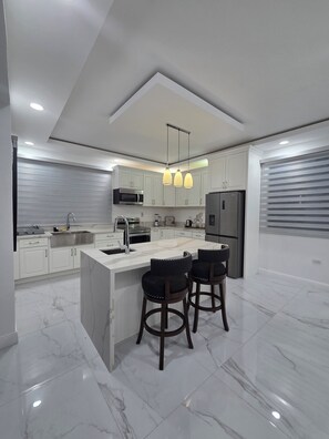 A modern kitchen with a central island, white cabinetry, stainless steel appliances, and pendant lighting. - Charisma Airbnb (Kingston)