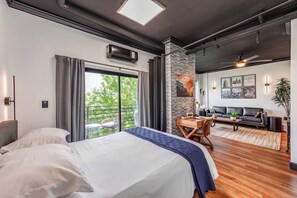 A modern bedroom with a large bed, a balcony, a dining area, and a living room with a sofa and a plant. - 4th Ward Industrial in the Heart of Charlotte - Apartment (Charlotte)