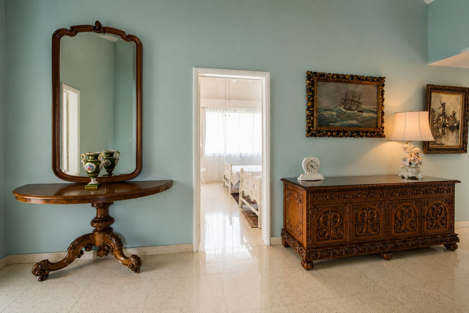 A room with a large mirror, a wooden side table with decorative items, a cabinet with intricate carvings, and a door leading to another room.