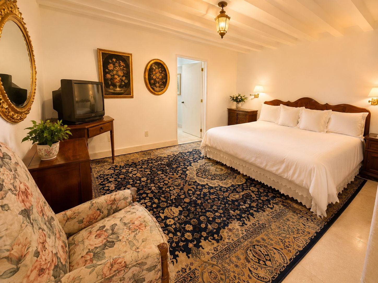 Una habitación de hotel con una cama grande, un televisor, un sillón tapizado con motivos florales, un alfombra estampado y dos lámparas fijadas a la pared.