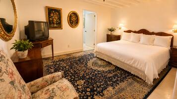 Una habitación de hotel con una cama grande, un televisor, un sillón tapizado con motivos florales, un alfombra estampado y dos lámparas fijadas a la pared.