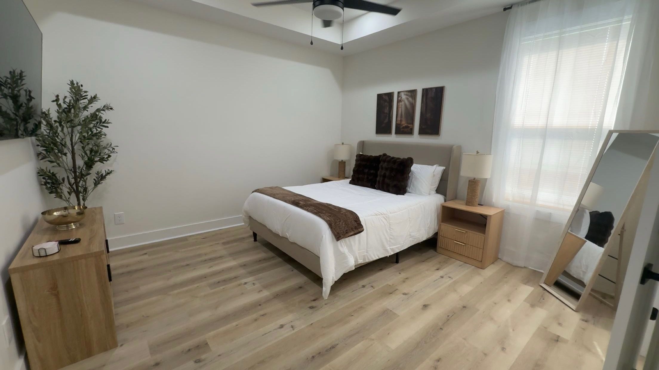 Une chambre à coucher comprenant un lit, des tables de chevet, un ventilateur de plafond, une fenêtre avec des rideaux et un miroir.