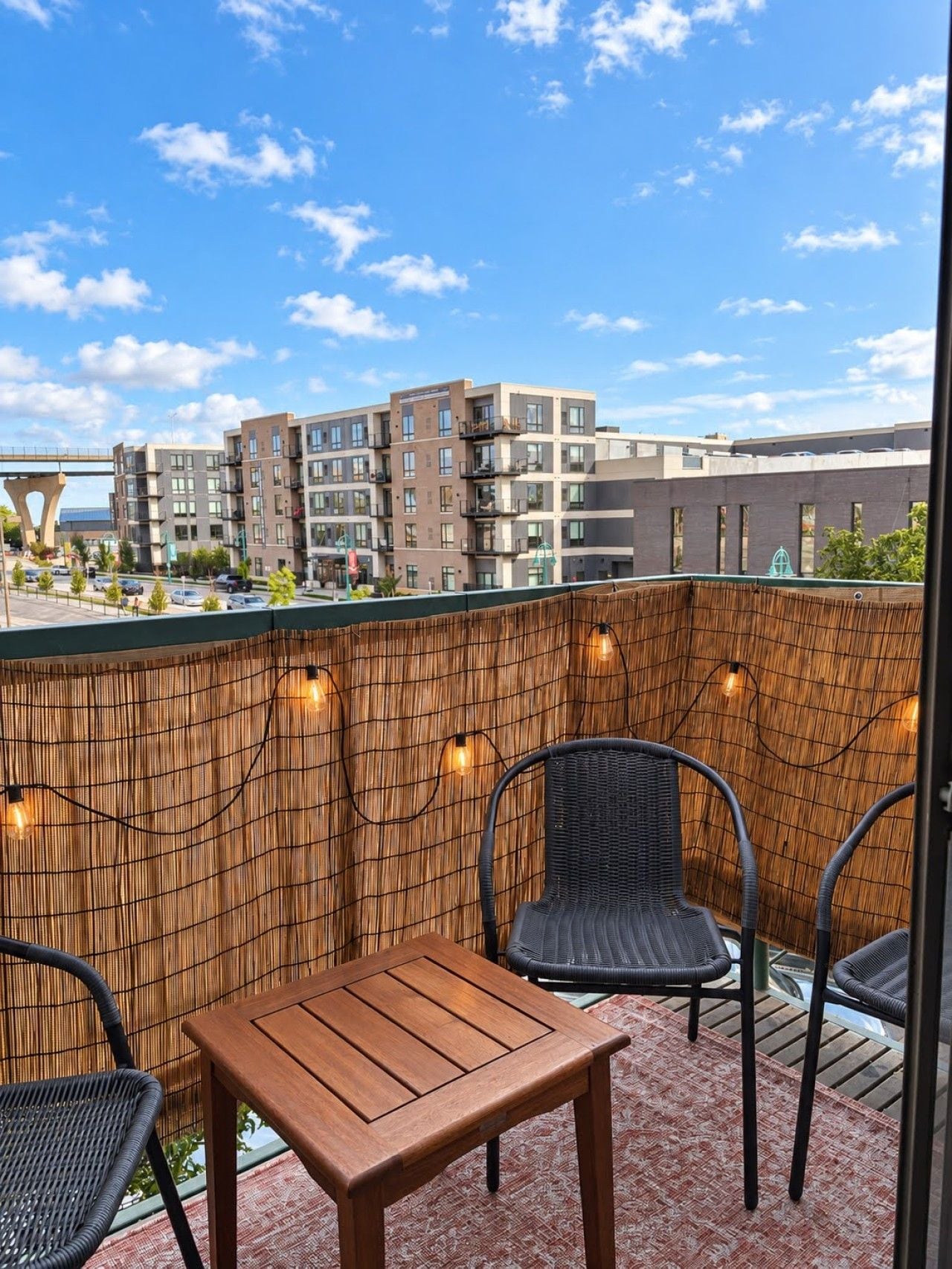 A balcony with a wooden table and two chairs, a bamboo wall, and string lights.