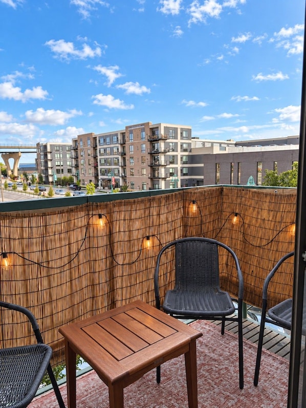 A balcony with a wooden table and two chairs, a bamboo wall, and string lights. - Third Ward Designer 2BR with Balcony | Sleeps 10 (Milwaukee)