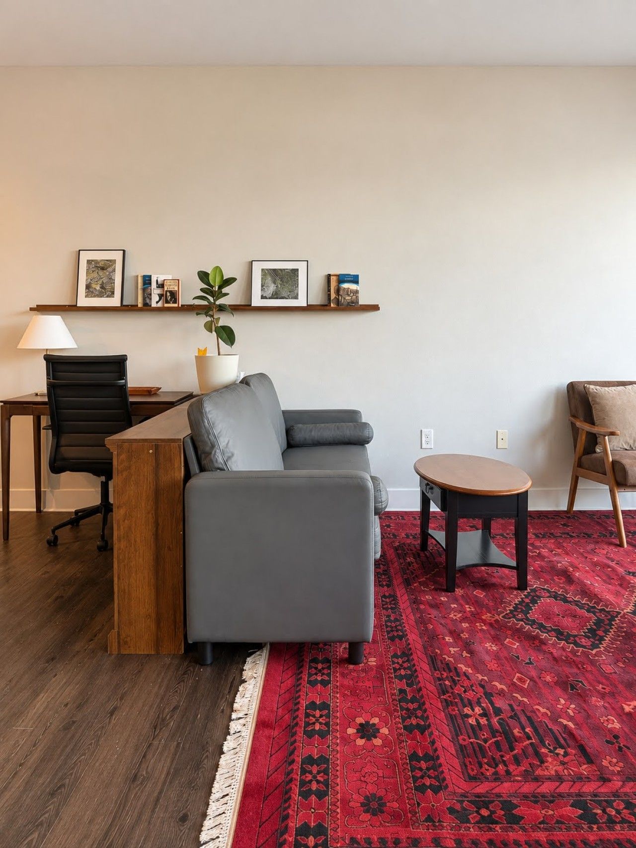 A modern living room with a grey sofa, a wooden coffee table, a red patterned rug, a black office chair, and a desk with a lamp.