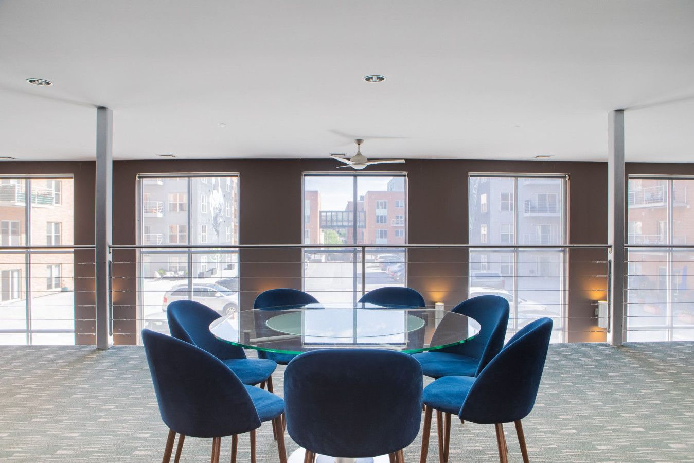 A conference room with a glass table surrounded by blue chairs, large windows with a city view, and a ceiling fan.