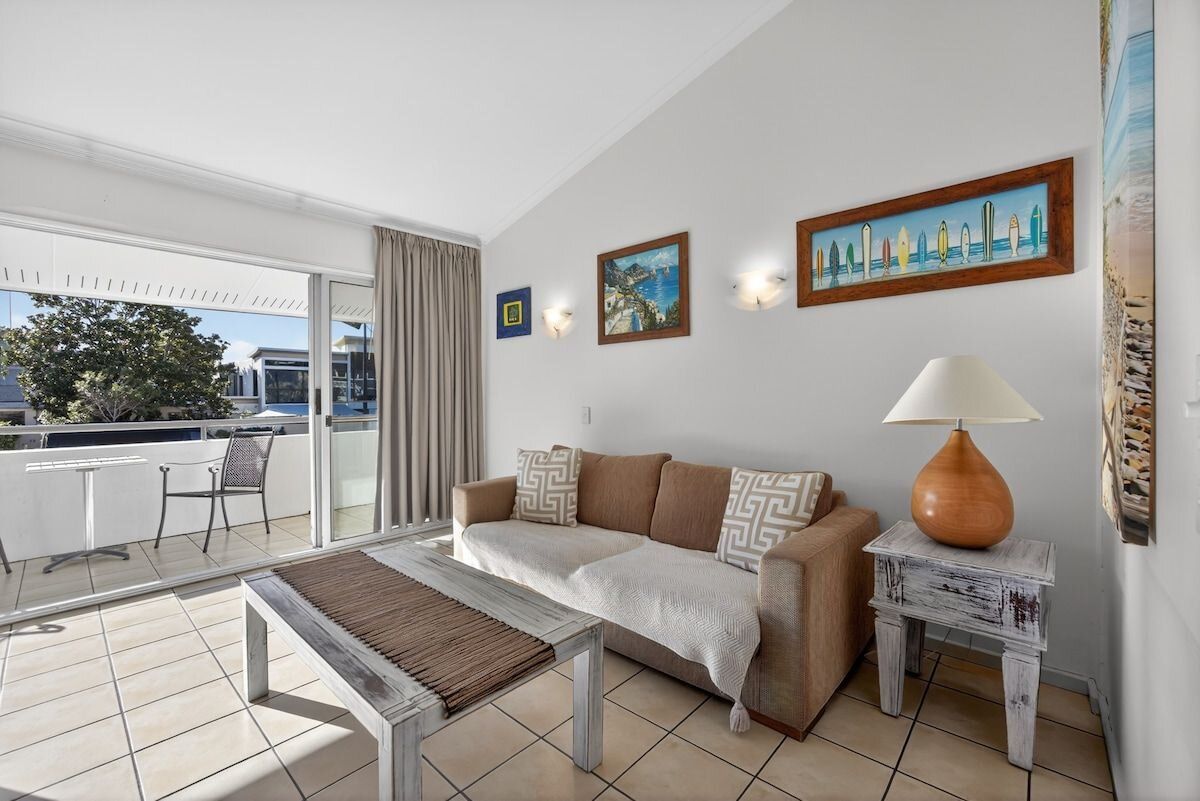 A living room with a sofa, a coffee table, and a lamp. There is a balcony with a view of the outside.