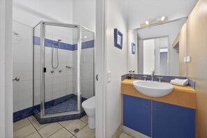 A bathroom with a glass shower enclosure, a white toilet, a sink with a blue countertop, and a mirror above the sink. - Couples Budget Stay on Noosa Parade! (Noosa Heads)