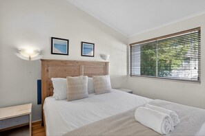 Quarto com cabeceira de madeira, cama com roupa de cama branca, duas luminárias fixadas na parede e uma janela com persianas.
