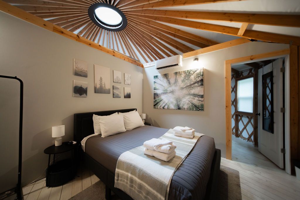 A bedroom with a bed, a nightstand, a lamp, a large window, and a door leading to another room.
