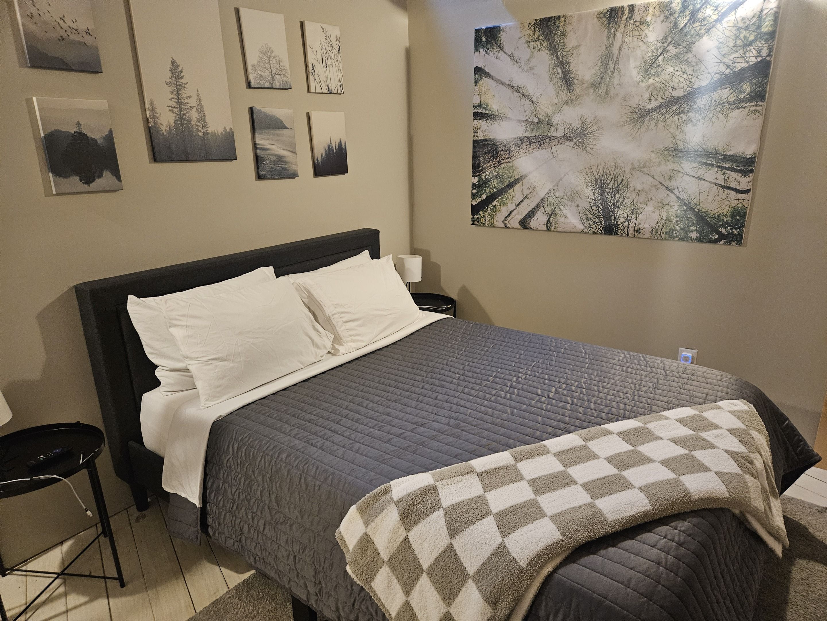 Ein ordentlich bezogenes Bett mit einer grauen Tagesdecke und einer karierten Zudecke, ein Nachttisch mit einer Lampe und eine Wand mit gerahmten Bildern zum Thema Natur.