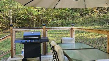 Terraza exterior con mesa y sillas de vidrio, una parrilla y un gran paraguas que proporciona sombra.