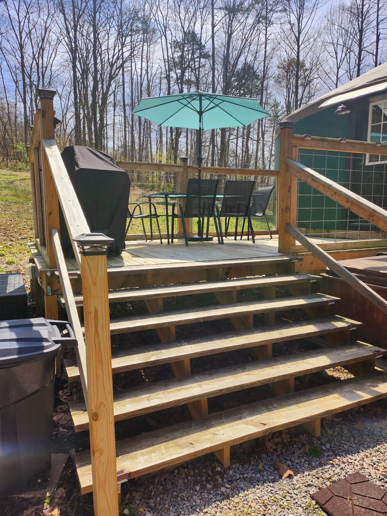 Des marches extérieures en bois mènent à une terrasse aménagée avec un barbecue, des chaises et un parasol.