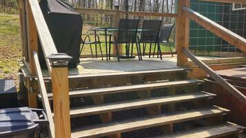 Des marches extérieures en bois mènent à une terrasse aménagée avec un barbecue, des chaises et un parasol.