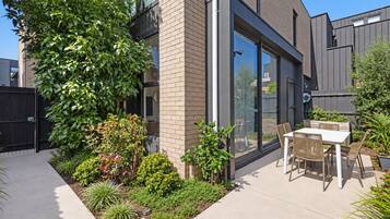 A modern patio with a dining table and chairs, surrounded by a variety of plants and a brick wall.
