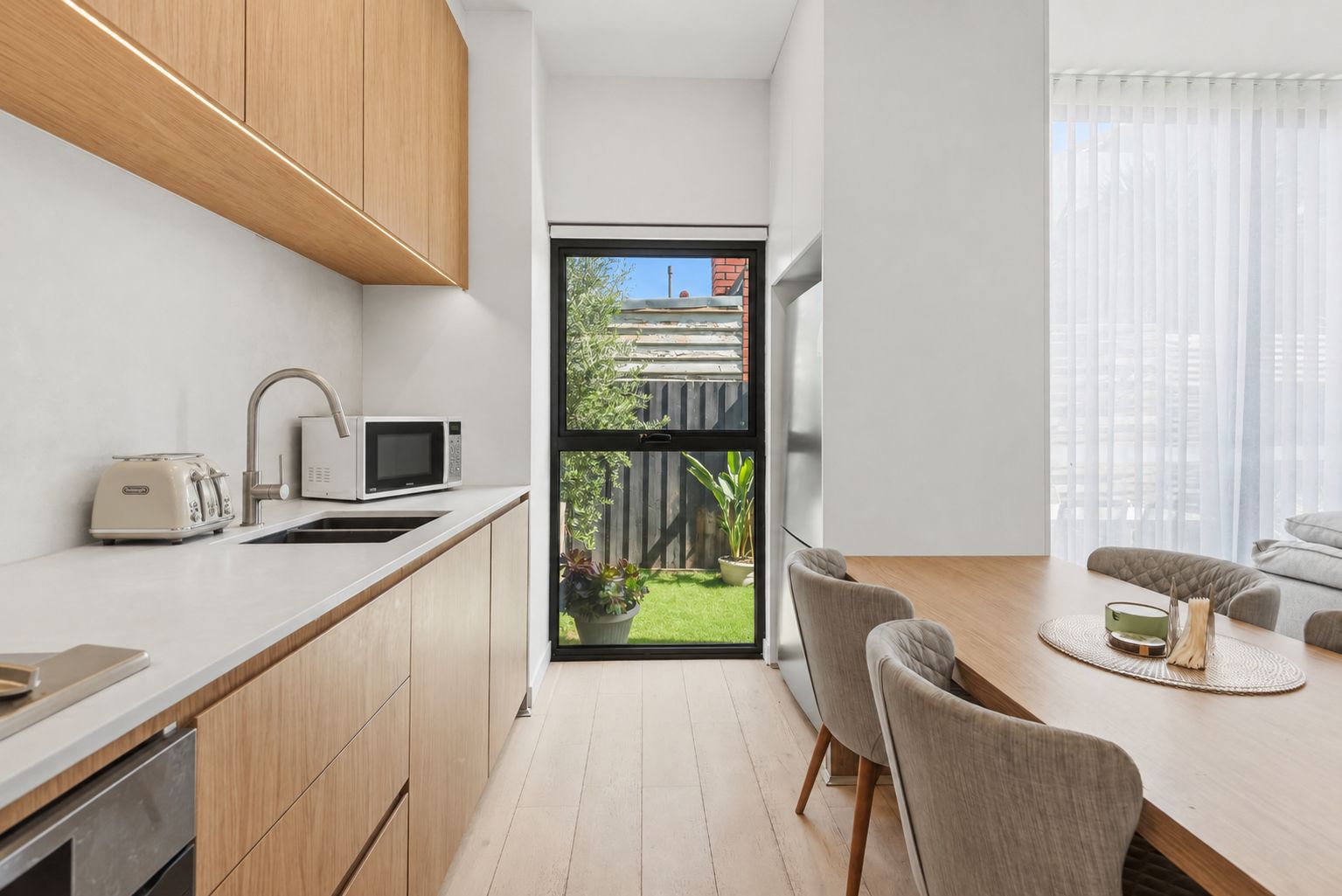 A modern kitchen with wooden cabinets, a microwave, and a toaster. There is a dining area with a table and chairs, and a view of a backyard through a large window.