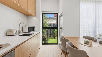 A modern kitchen with wooden cabinets, a microwave, and a toaster. There is a dining area with a table and chairs, and a view of a backyard through a large window.