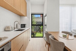 Een moderne keuken met houten kastjes, een magnetron en een broodrooster. Er is een eethoek met een tafel en stoelen, en een uitzicht op een achtertuin door een groot raam.