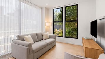 A modern living room with a grey sofa, a wooden side table, a flat-screen TV, and large windows with a view of greenery.