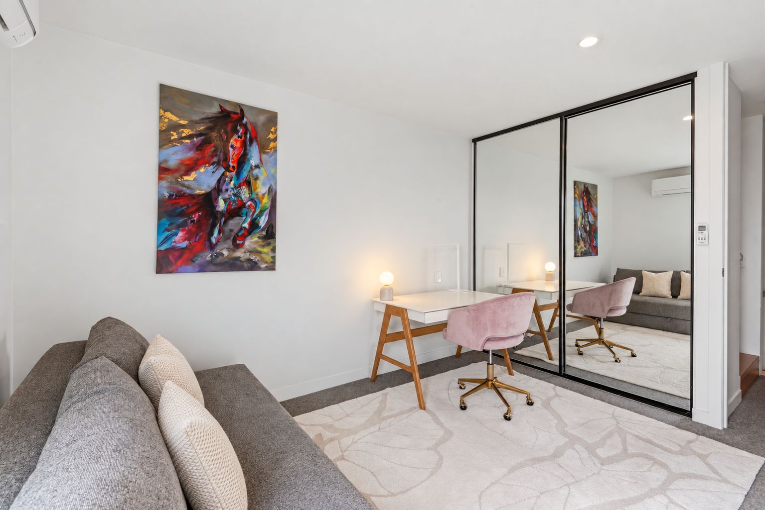 A modern room with a white desk, a pink chair, a large mirror, and a sofa with multiple cushions.