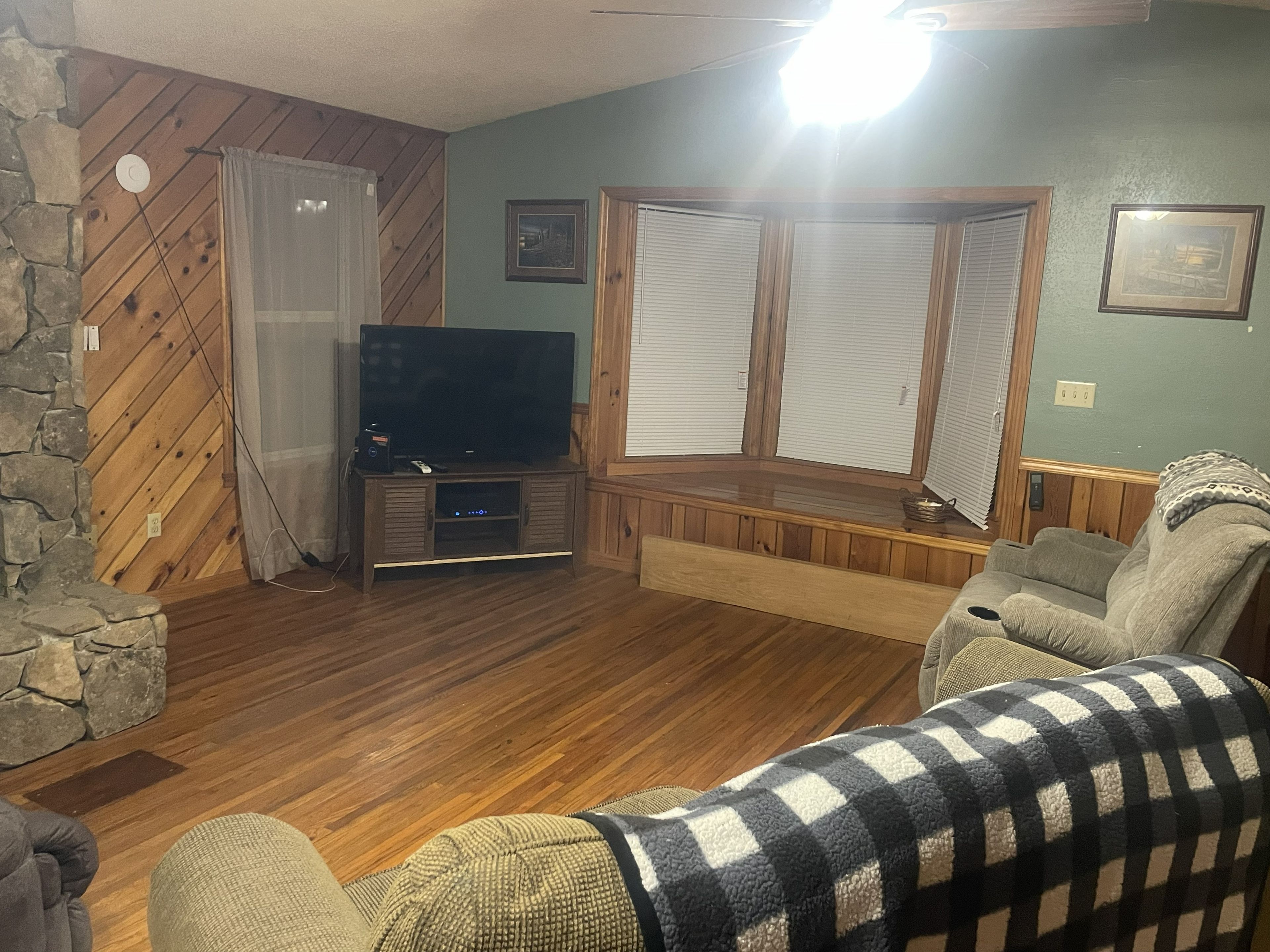 A living room with a television, a stone wall, wooden flooring, and a window with blinds.