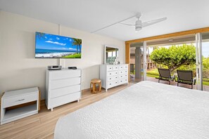 A bedroom with a large bed, a television, a dresser, a small stool, and a view of a balcony with chairs. - Kaanapali Maui Eldorado D103 Garden View 2 BD (Lahaina)