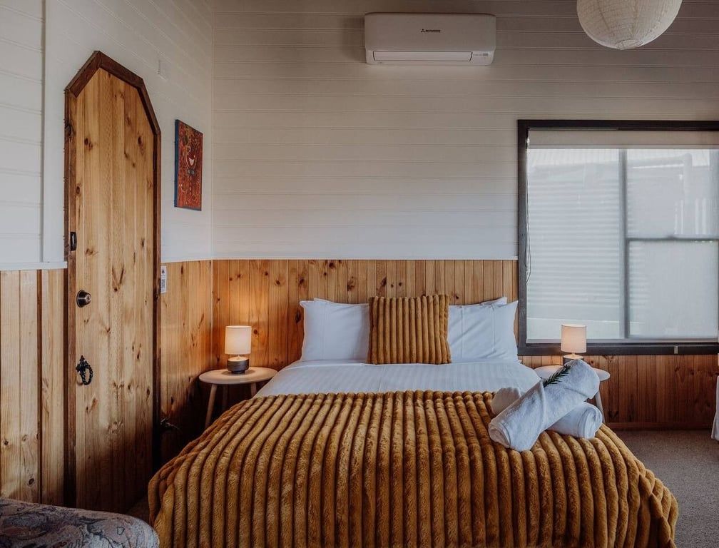 A hotel room with a wooden headboard, a bed with a striped blanket, two bedside lamps, and a window with blinds.