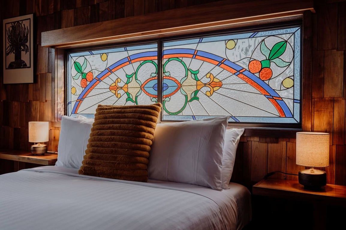 A hotel room with a bed, two bedside lamps, a large stained glass window, and a wooden headboard.