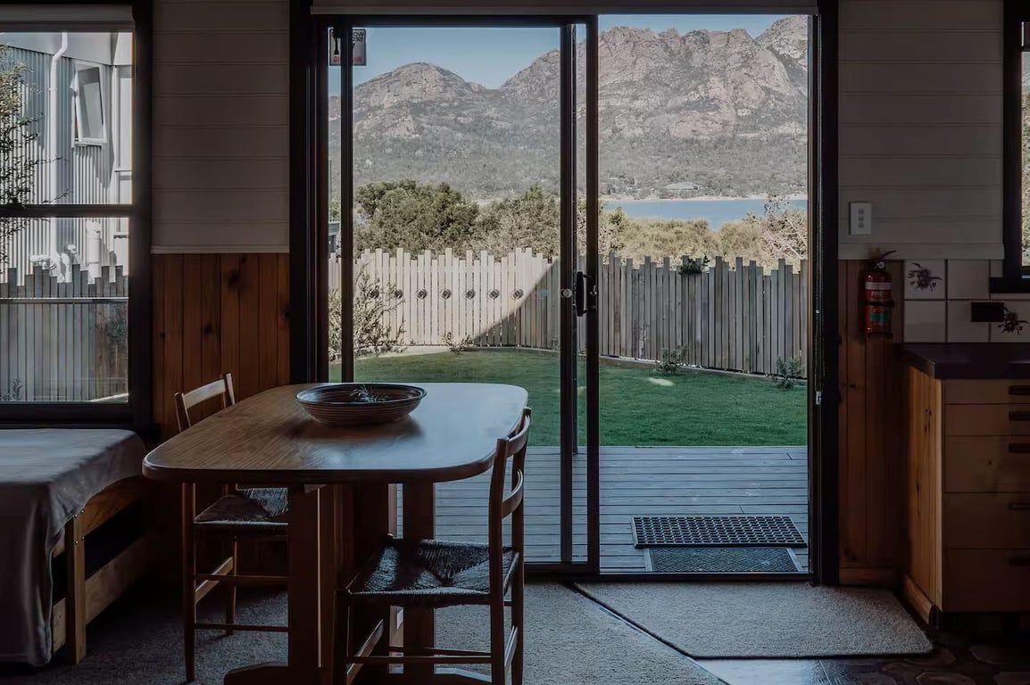 Een kamer met een houten tafel en stoelen, een schuifdeur naar een achtertuin met een houten omheining en bergen op de achtergrond.