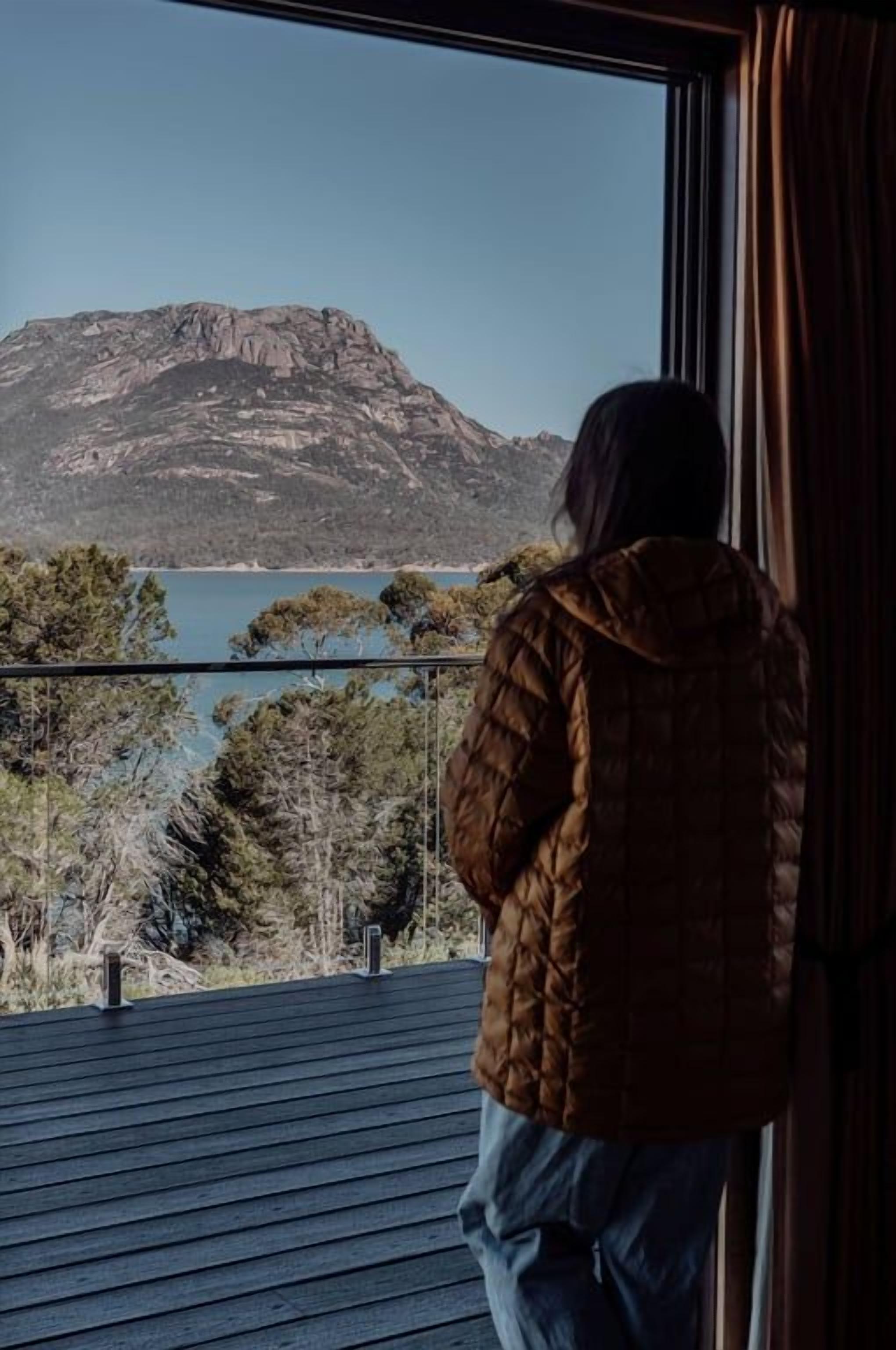 A person standing on a balcony overlooking a mountainous landscape and a body of water.