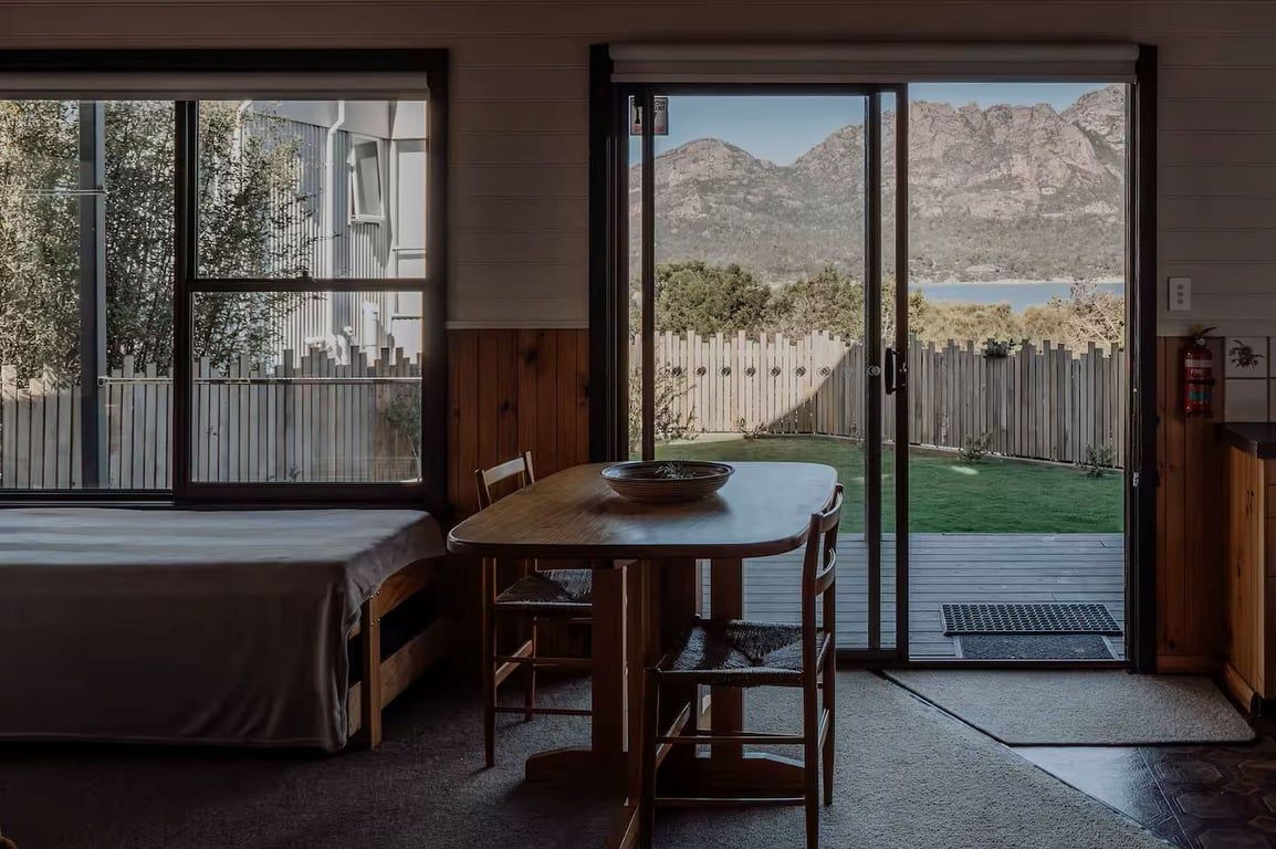 A bedroom with a bed, a dining table with chairs, and a sliding glass door leading to a backyard with a view of mountains and a lake.