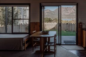 A bedroom with a bed, a dining table with chairs, and a sliding glass door leading to a backyard with a view of mountains and a lake.