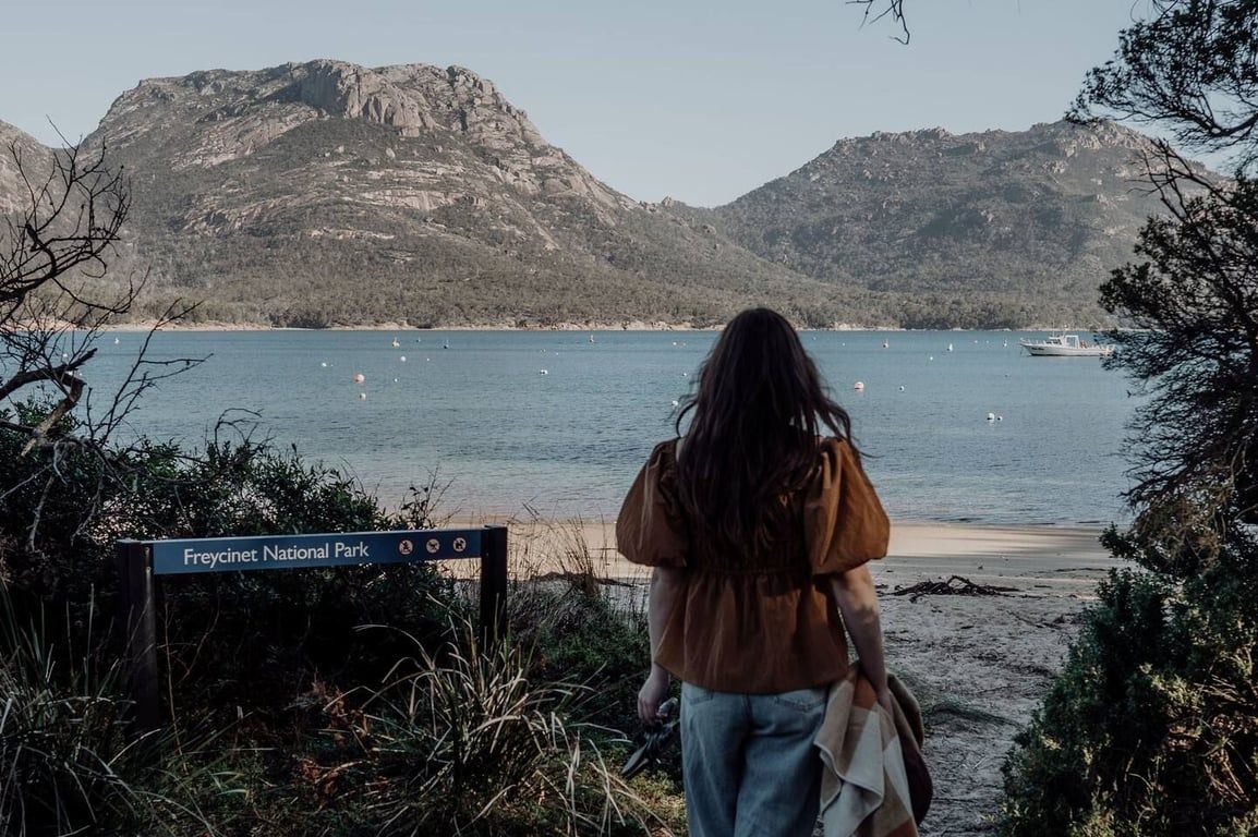 A person standing at the edge of a beach with a sign indicating Freycinet National Park.