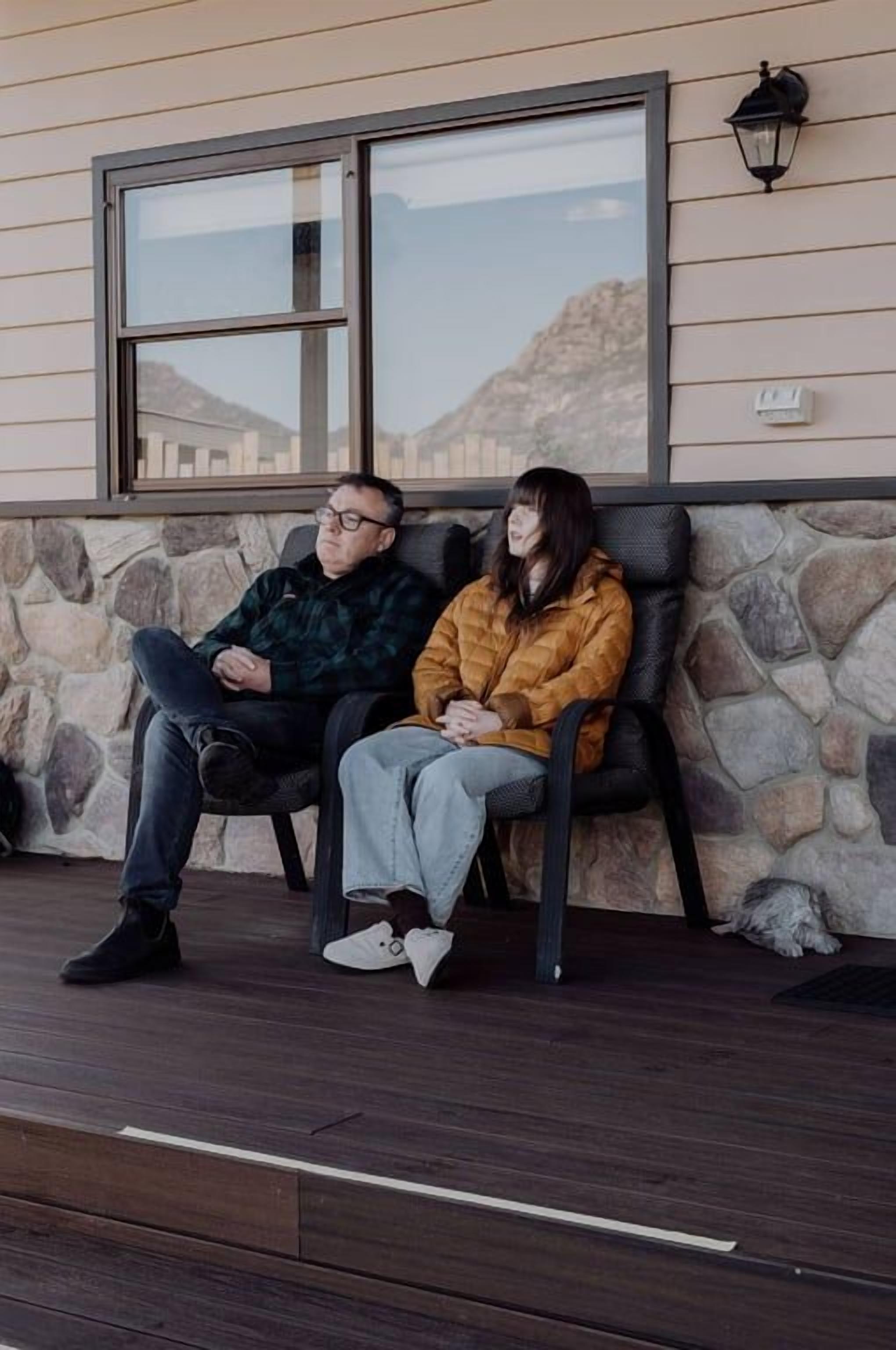 Two people sitting on a bench outside a building with stone walls and a window.