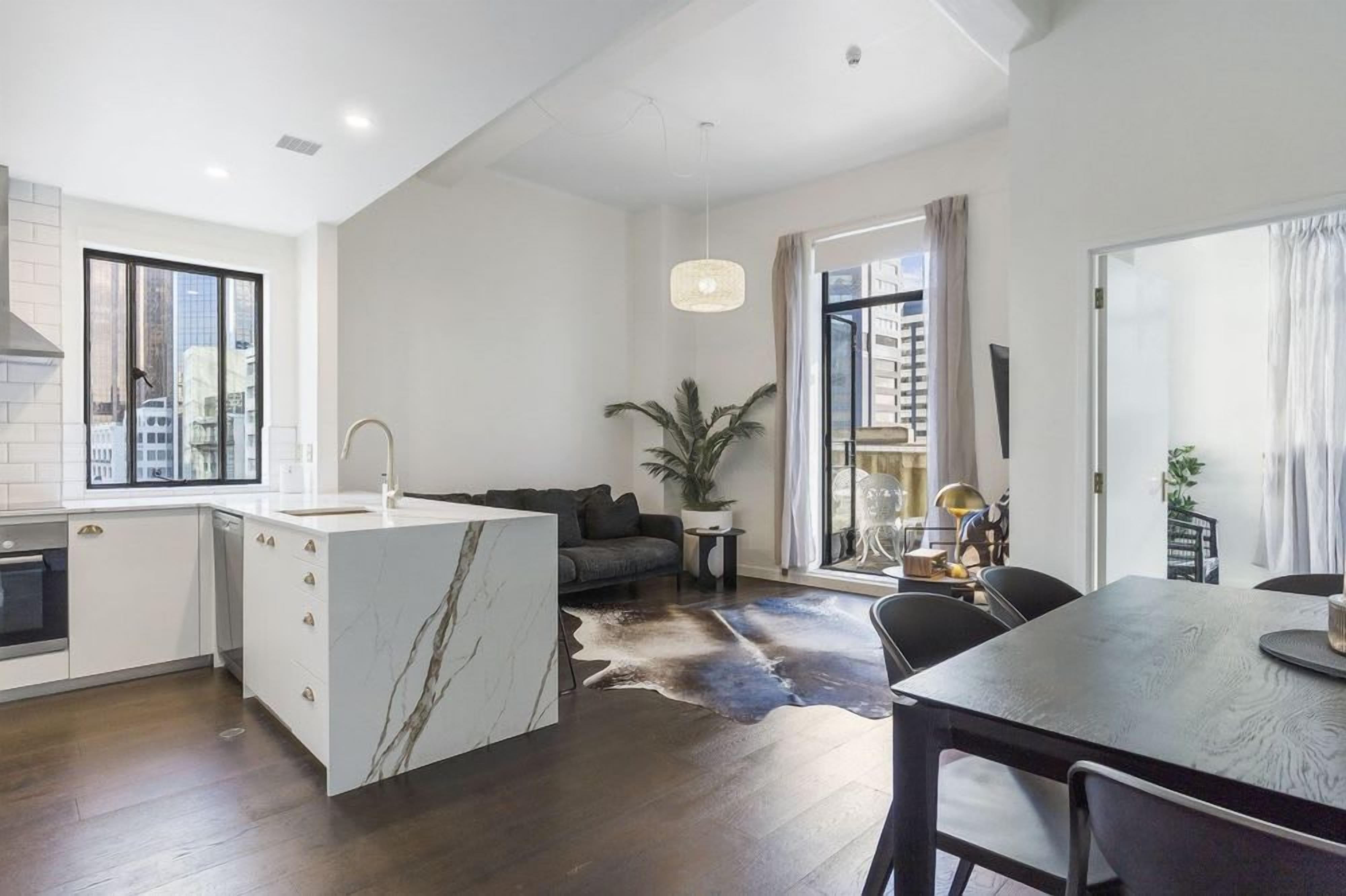 A modern kitchen with a marble island, a dining area with a table and chairs, a sofa, and a large window with a city view.