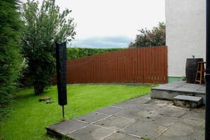 A paved patio area with a wooden fence and a grassy lawn. - Bright 4BR Cottage | Enclosed Garden & Parking (Ballycastle)