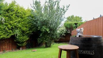 A wooden barrel labeled 'BUSHMILLS' in a backyard with a wooden stool, a tree, and a fence.