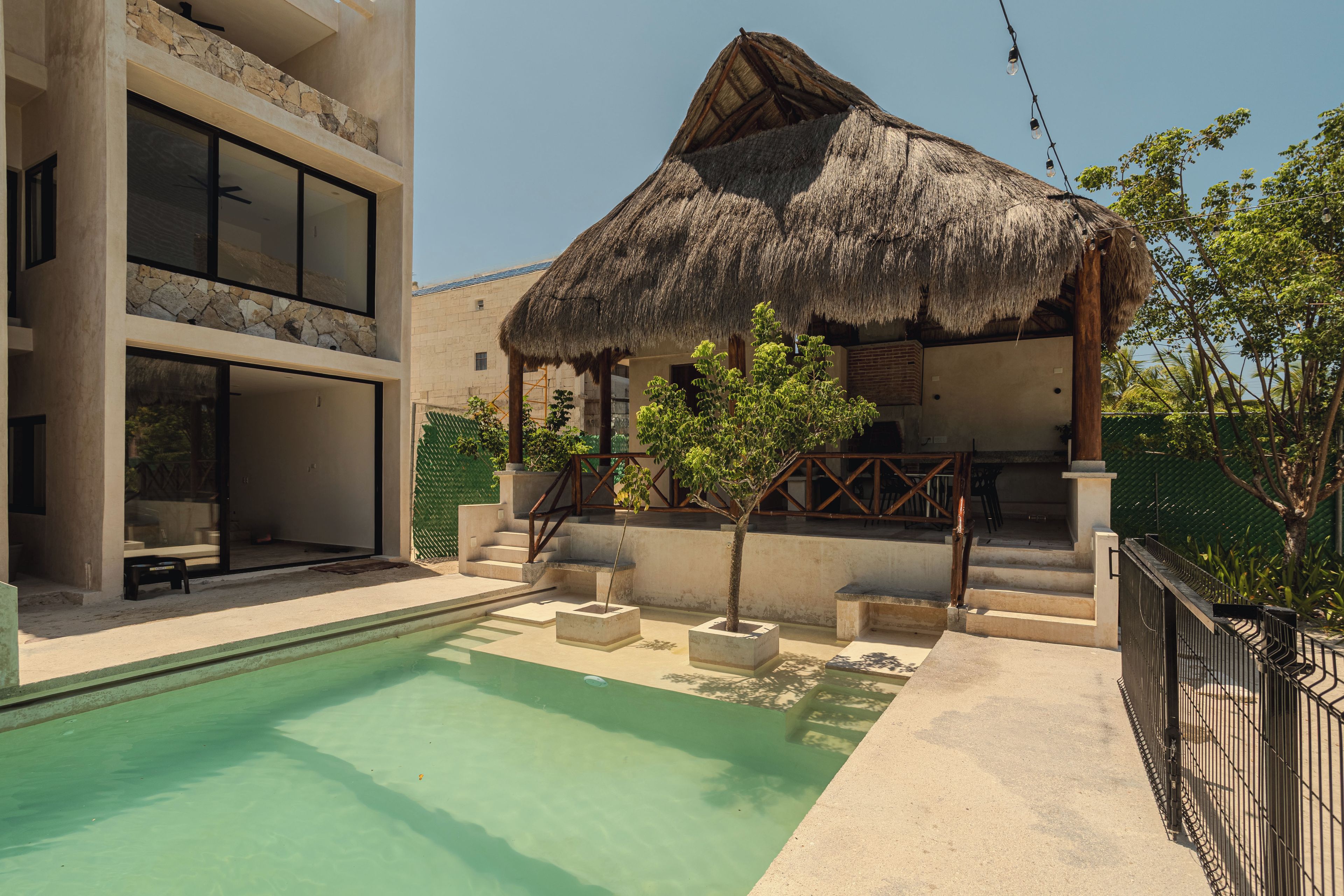 Un área de piscina con una estructura de techo de paja, un edificio de varios pisos y un jardín cercado.