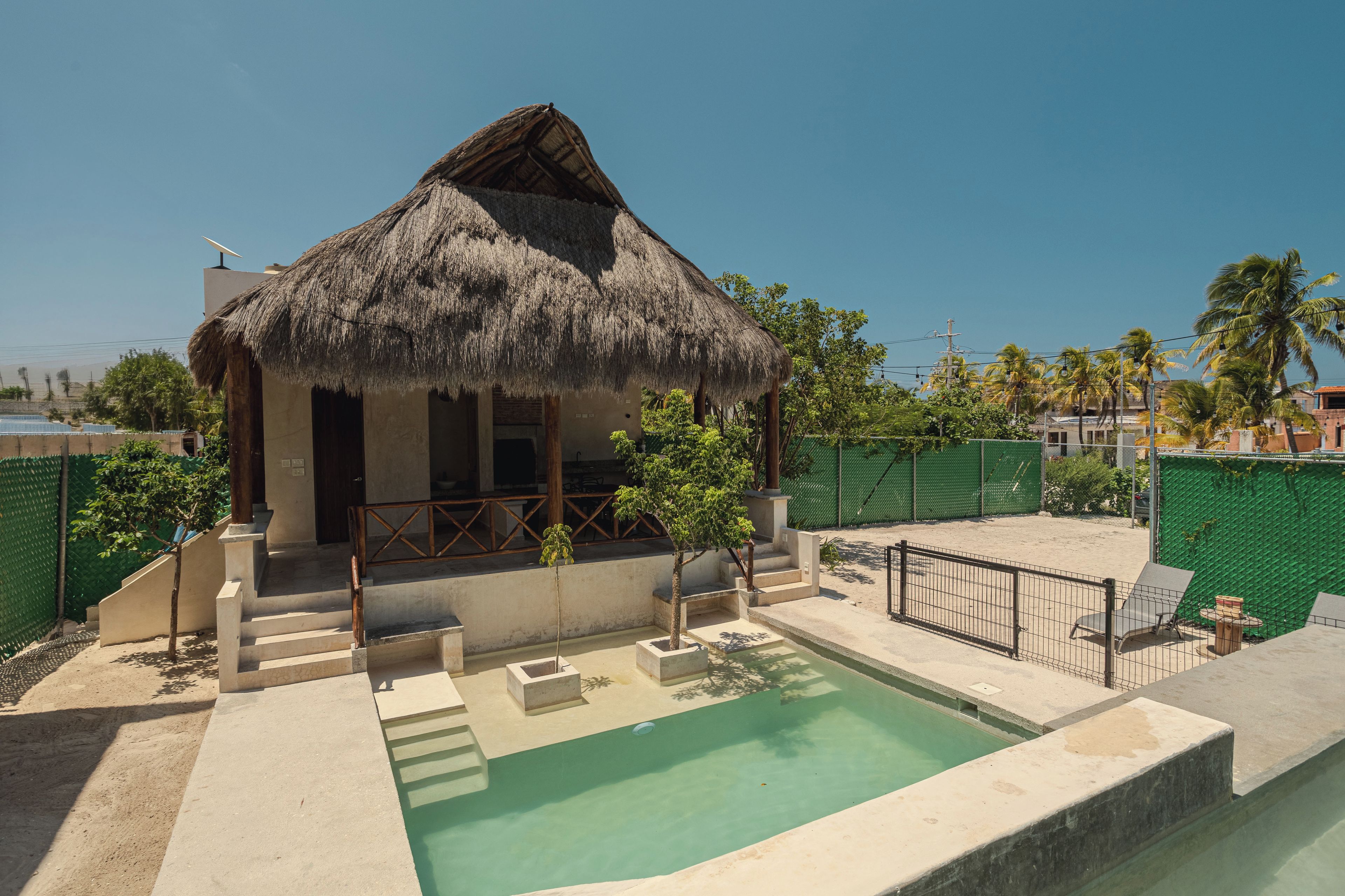 A thatched-roof bungalow with a swimming pool, surrounded by greenery and a fenced area.